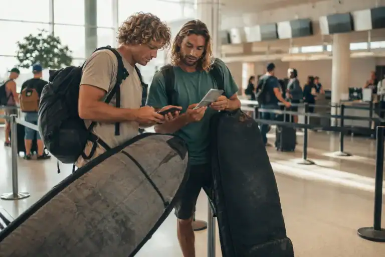 Two surfers reviewing travel documents with board bags at an airport